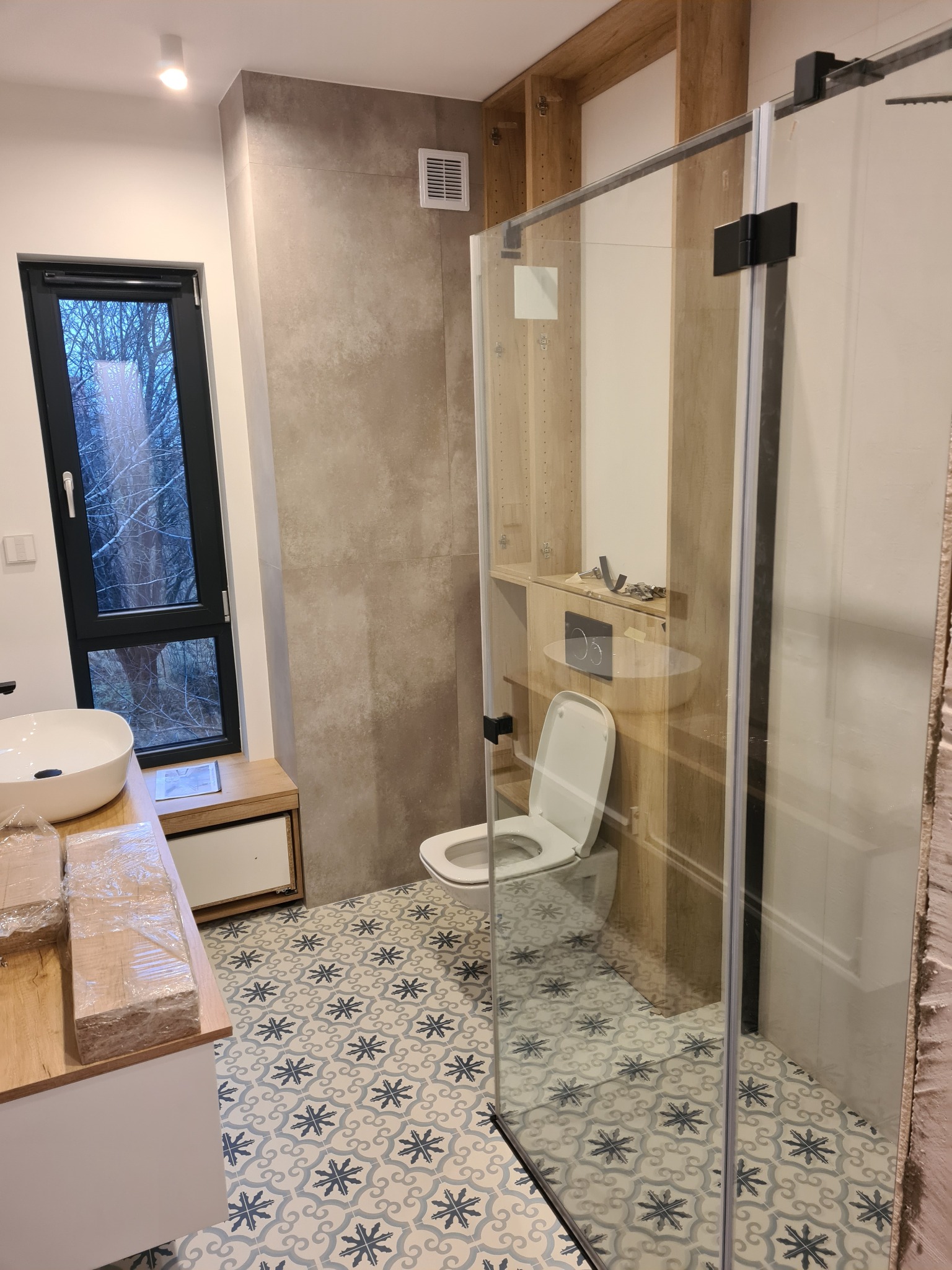 Nowoczesna łazienka w trakcie wykańczania: kabina prysznicowa ze szkła hartowanego, umywalka nablatowa, toaleta podwieszana i wzorzysta podłoga, widoczne narzędzia na półce.