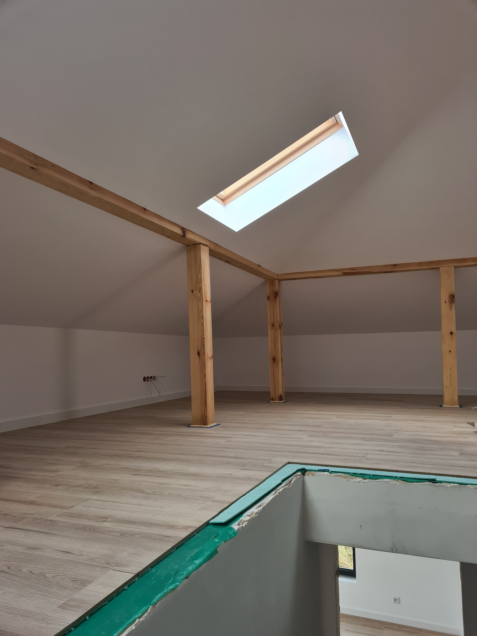 Poddasze po remoncie: drewniane belki konstrukcyjne, wykończona podłoga z paneli, okno dachowe, widoczne kable elektryczne w ścianie, przygotowane do dalszych prac wykończeniowych.