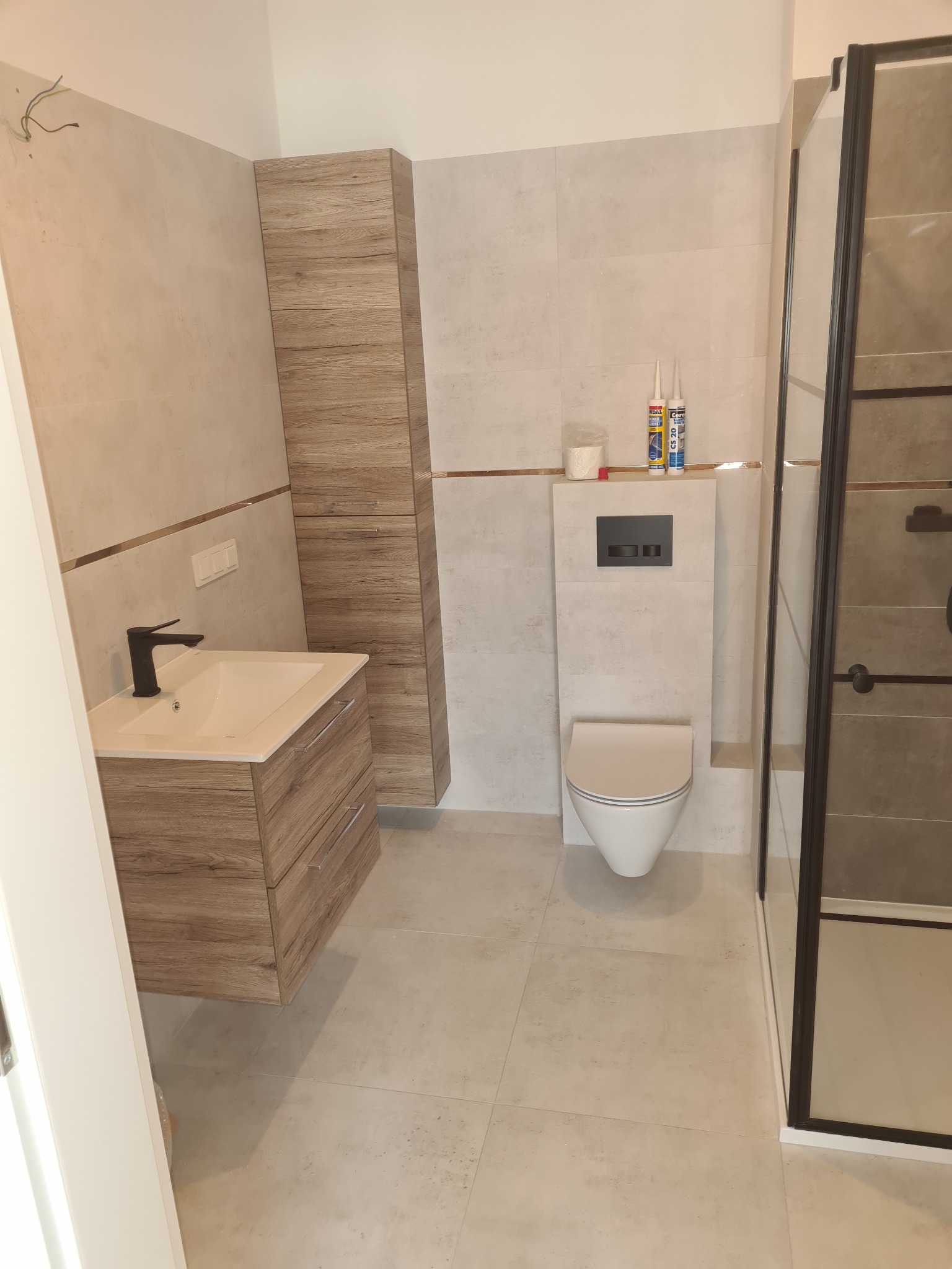 Nowoczesna łazienka z drewnopodobną szafką pod umywalką z czarną baterią, wiszącą toaletą i kabiną prysznicową z czarnymi profilami, wykończona beżowymi płytkami.