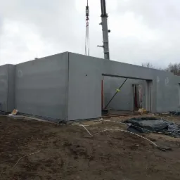 Montaż prefabrykowanych ścian domu jednorodzinnego za pomocą dźwigu, widoczne otwory okienne i drzwiowe, teren budowy w trakcie prac.