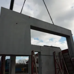 Montaż prefabrykowanego elementu betonowego z otworem okiennym za pomocą dźwigu na placu budowy, widoczne drabiny i inne elementy konstrukcyjne.