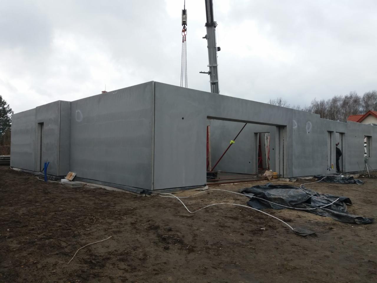 Montaż prefabrykowanych ścian domu jednorodzinnego za pomocą dźwigu, widoczne otwory okienne i drzwiowe, teren budowy w trakcie prac.