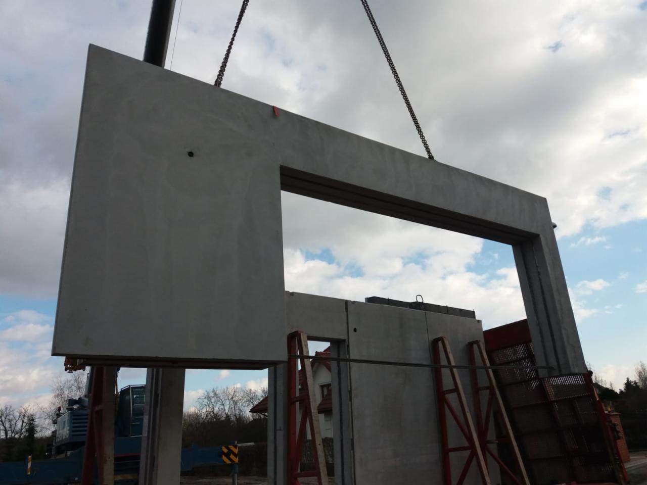 Montaż prefabrykowanego elementu betonowego z otworem okiennym za pomocą dźwigu na placu budowy, widoczne drabiny i inne elementy konstrukcyjne.