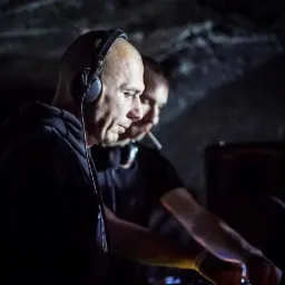 Dwóch mężczyzn w słuchawkach w ciemnym pomieszczeniu, jeden z papierosem w ustach, miksujący muzykę na konsoli didżejskiej.