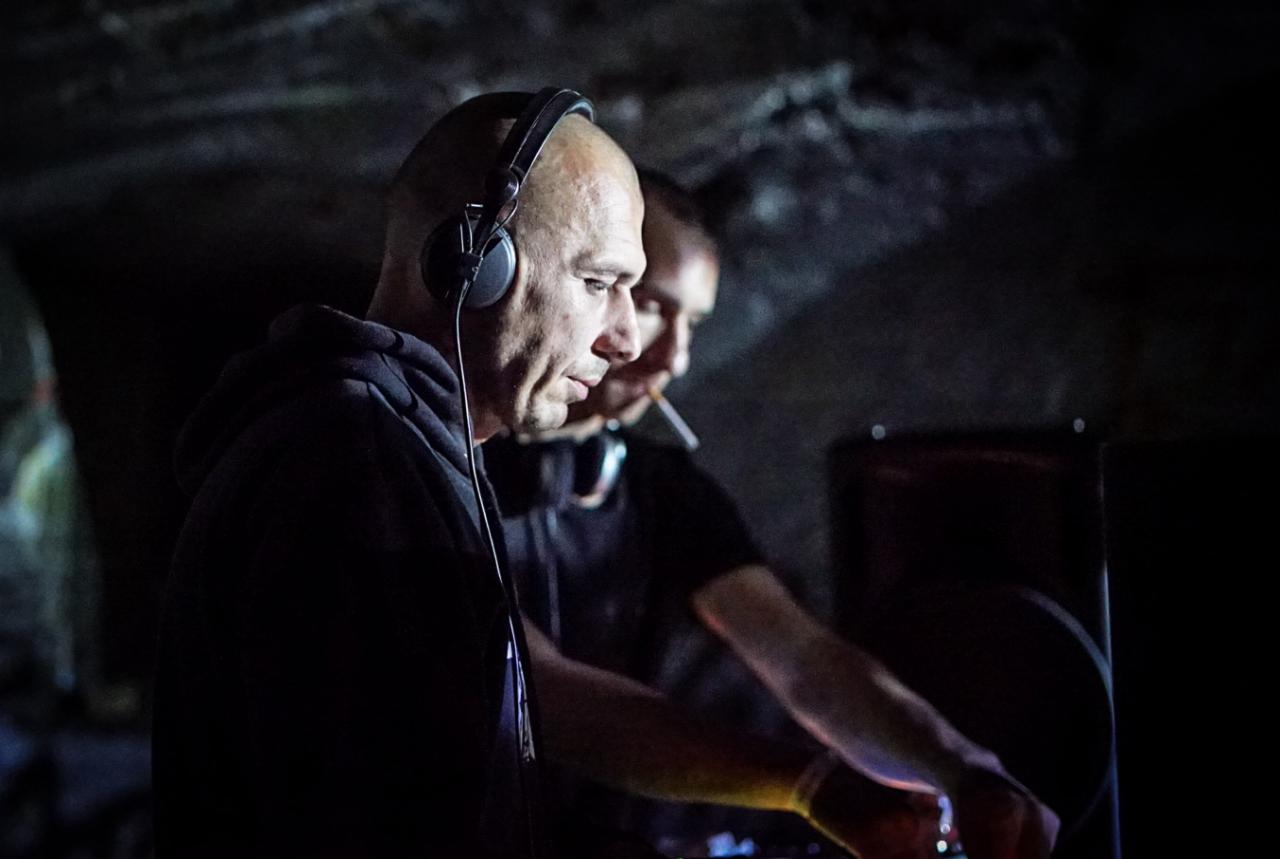 Dwóch mężczyzn w słuchawkach w ciemnym pomieszczeniu, jeden z papierosem w ustach, miksujący muzykę na konsoli didżejskiej.