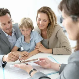 Rodzina z synem podpisuje dokument na tablecie, obsługiwana przez doradcę finansowego w jasnym biurze. Widoczne wykresy i dokumenty.