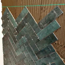 Układanie ciemnozielonych, prostokątnych płytek w jodełkę na ścianie, z widocznymi krzyżykami dystansowymi i poziomą linią lasera.