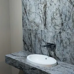 Umywalka nablatowa o owalnym kształcie na blacie z płytek imitujących marmur z wyraźnym, ciemnym żyłowaniem, zamontowana na tle ściany wyłożonej identycznymi płytkami, widoczny minimalistyczny kran...