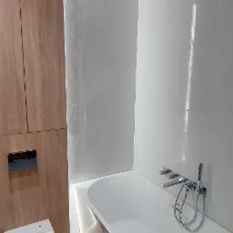 Nowoczesna łazienka z białą wanną, wiszącą toaletą i drewnianą zabudową, wykończona jasnymi płytkami z subtelną fakturą, oświetlona ledowym światłem.