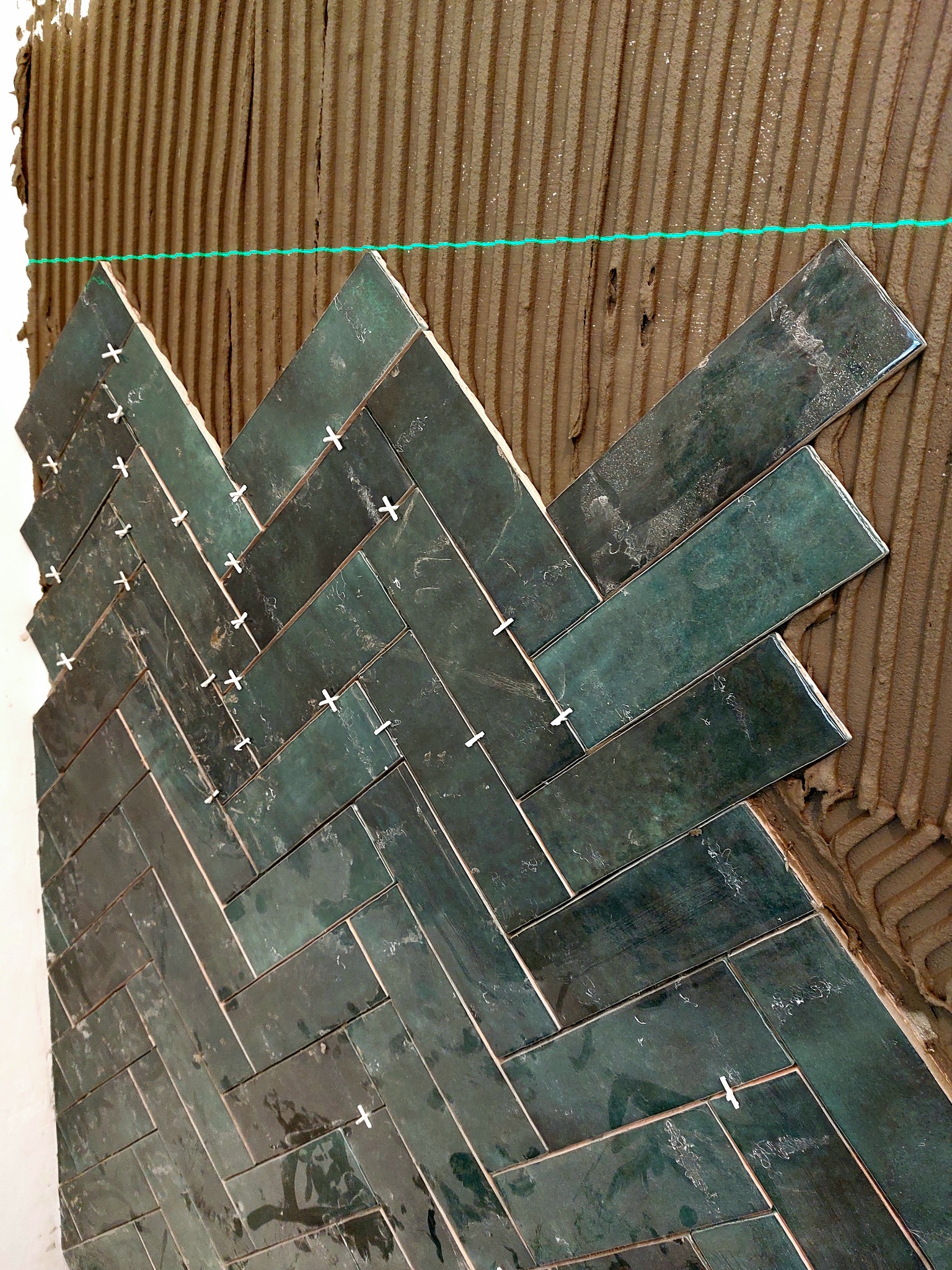 Układanie ciemnozielonych, prostokątnych płytek w jodełkę na ścianie, z widocznymi krzyżykami dystansowymi i poziomą linią lasera.