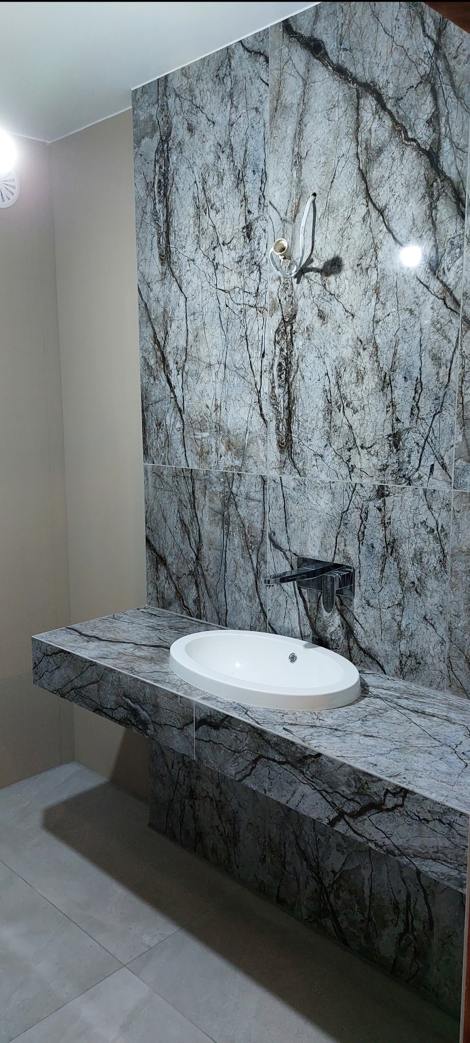 Umywalka nablatowa o owalnym kształcie na blacie z płytek imitujących marmur z wyraźnym, ciemnym żyłowaniem, zamontowana na tle ściany wyłożonej identycznymi płytkami, widoczny minimalistyczny kran...