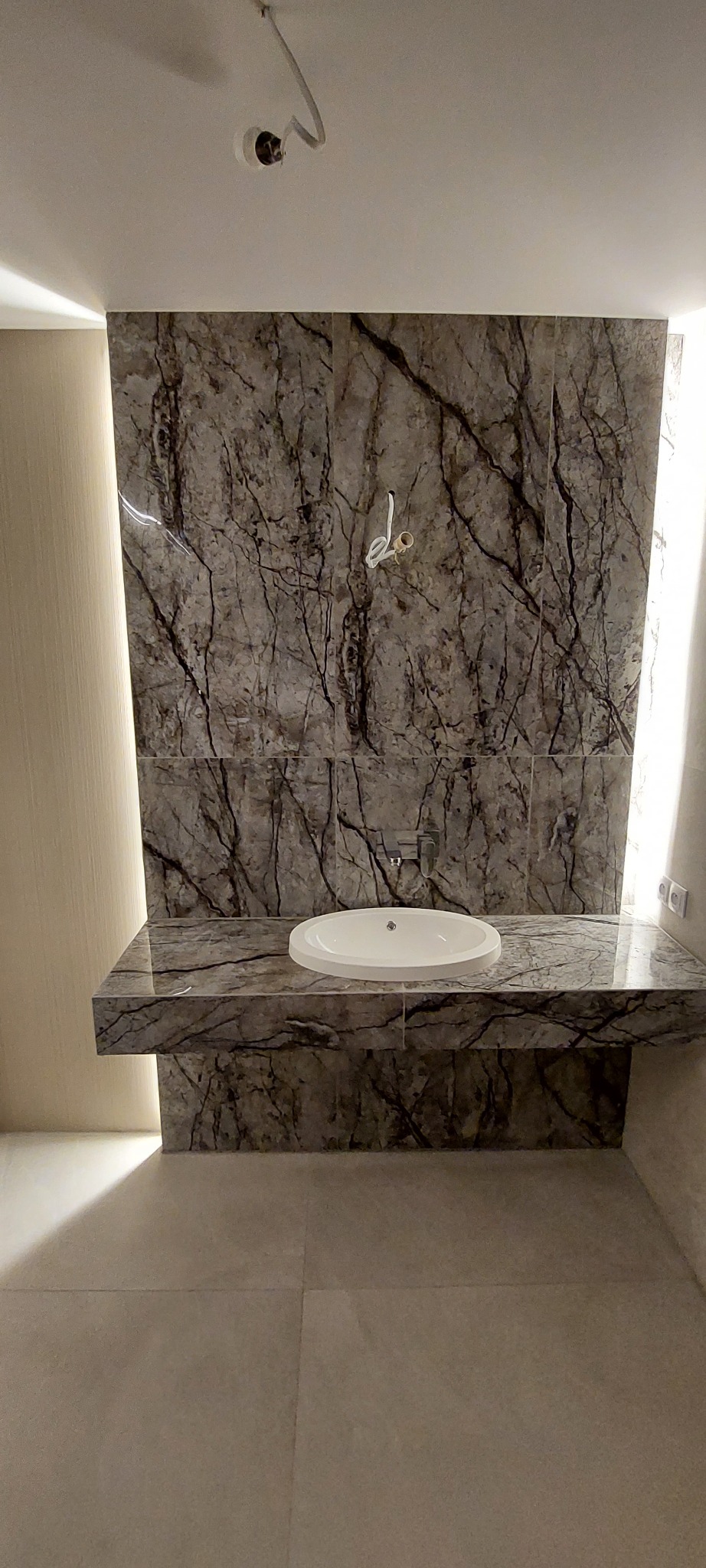 Łazienka w trakcie wykańczania: marmurowe płytki na ścianie i blacie pod umywalkę, widoczne wystające kable oświetleniowe, brakujące wykończenie sufitu.
