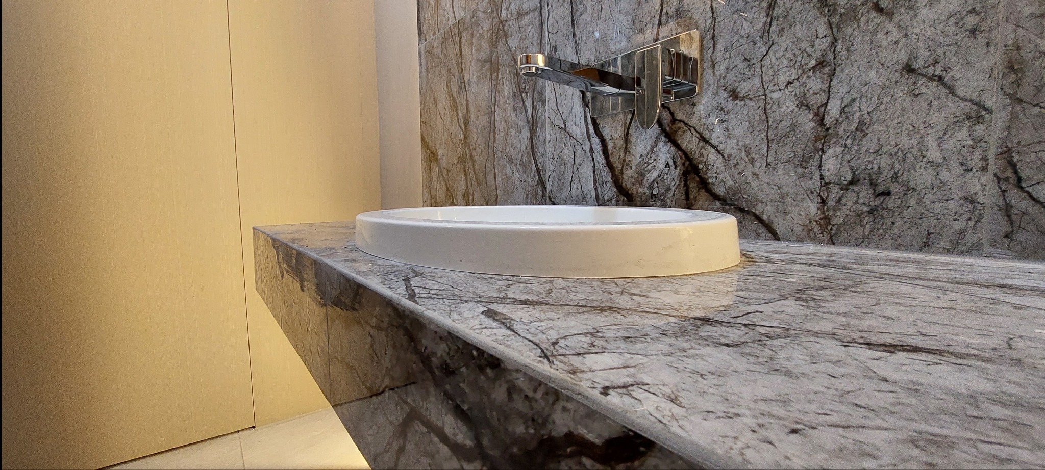 Biała, okrągła umywalka nablatowa na blacie z szarego marmuru z wyraźnym użyleniem, nowoczesna bateria chromowana zamontowana na ścianie pokrytej płytami imitującymi kamień.