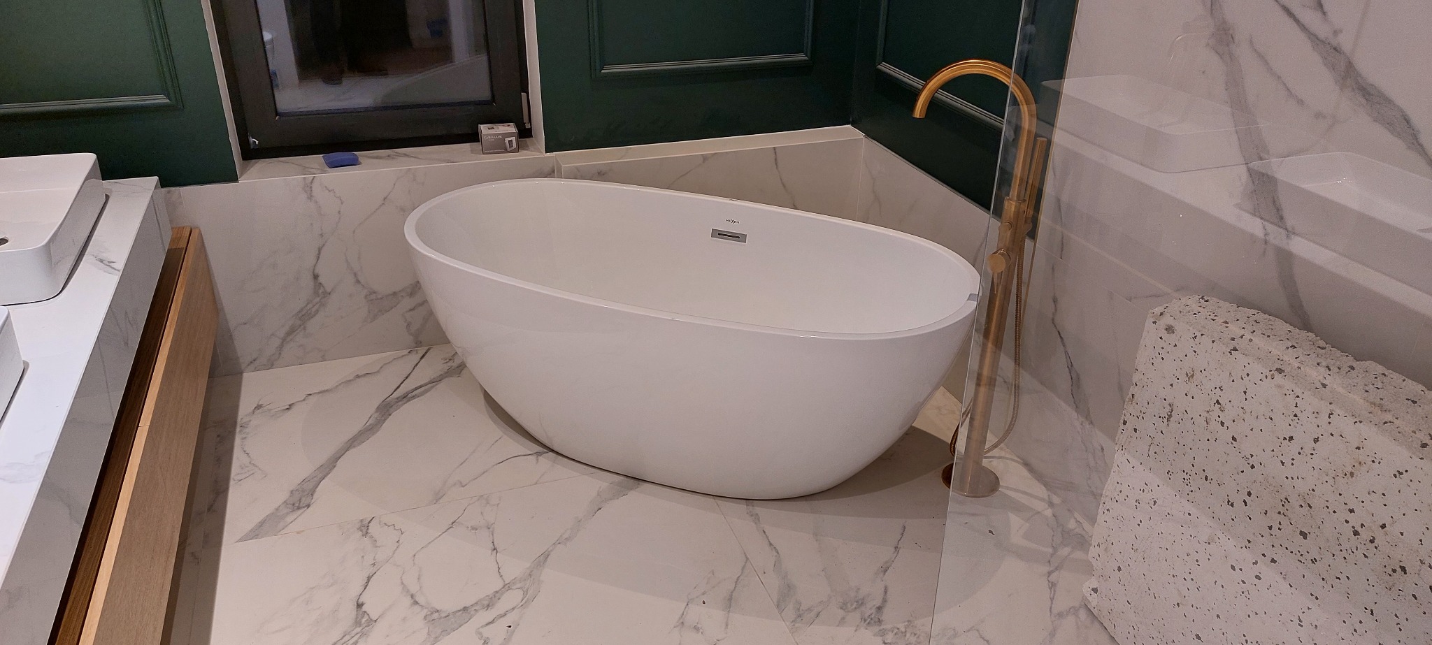 Wolnostojąca, owalna wanna z baterią podłogową w kolorze złotym, na tle marmurowych płytek i ciemnozielonej ściany z dekoracyjną sztukaterią.