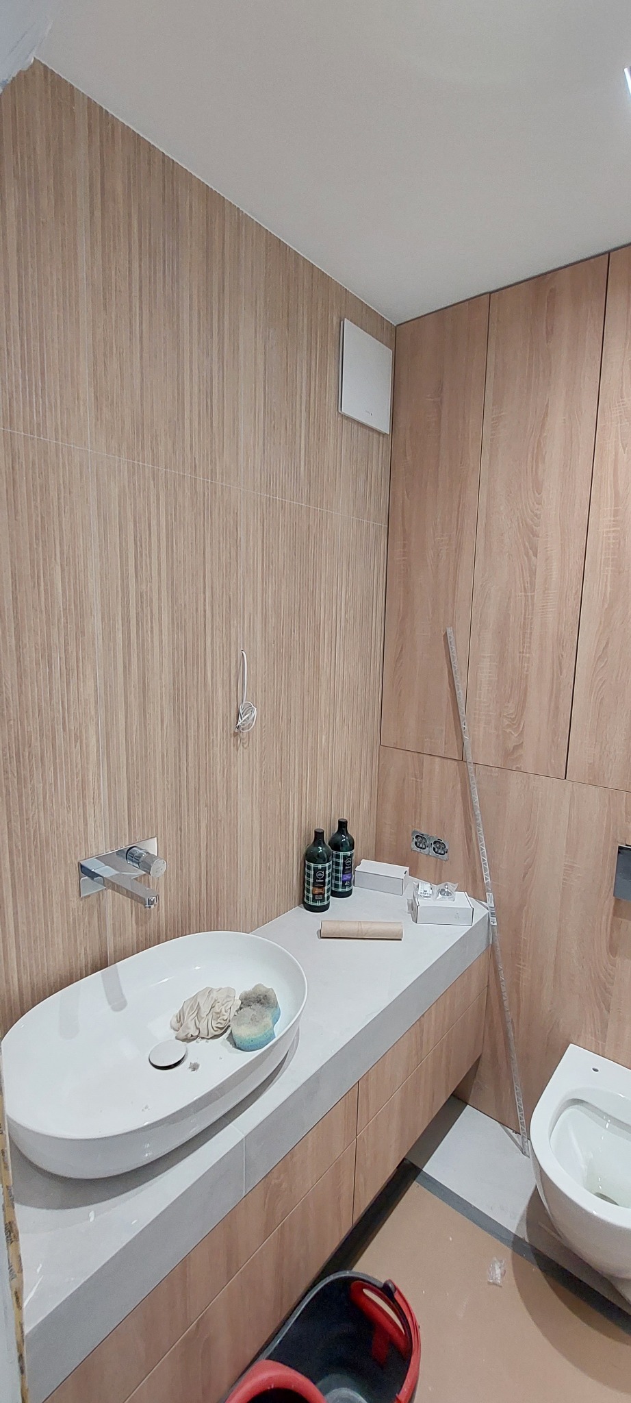 Łazienka w trakcie wykańczania: drewnopodobne płytki ścienne, umywalka nablatowa z chromowaną baterią, akcesoria i wiadro na podłodze.