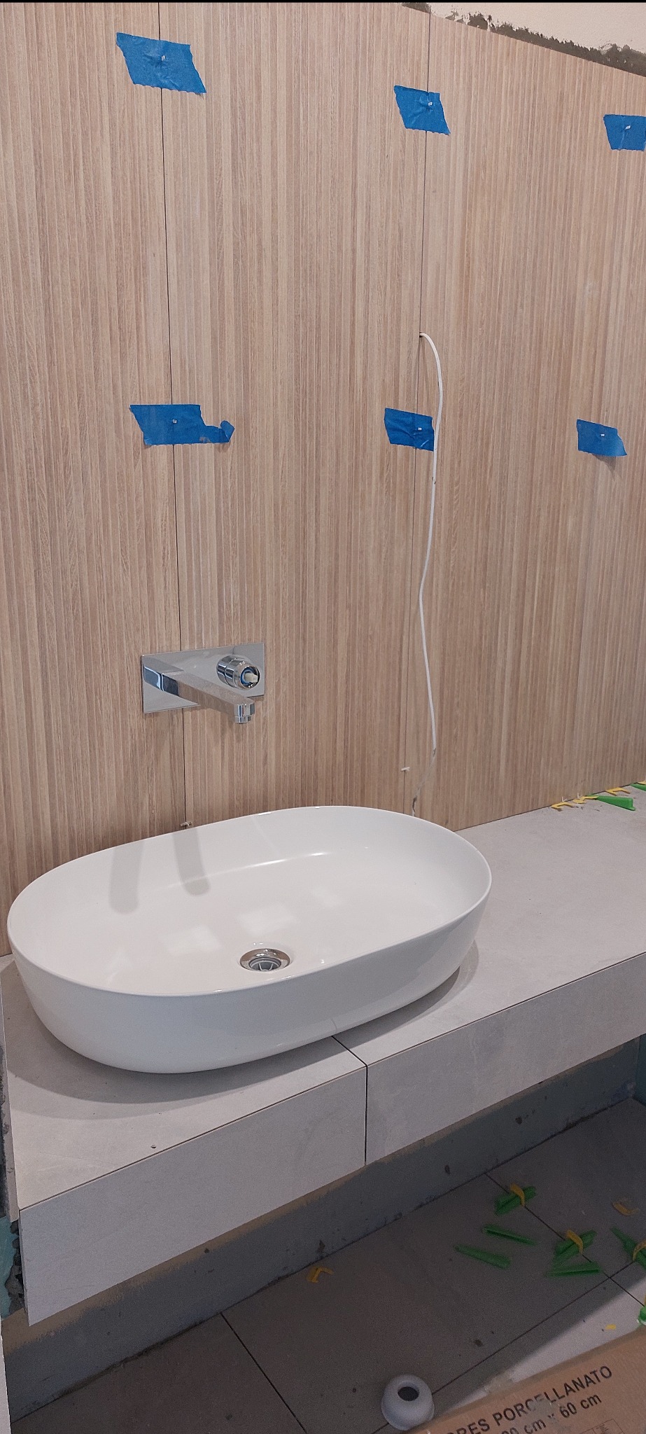 Montaż umywalki nablatowej na szarym blacie z płytek, widoczne płytki ścienne imitujące drewno z niebieską taśmą malarską i chromowana bateria podtynkowa, na podłodze krzyżyki dystansowe.