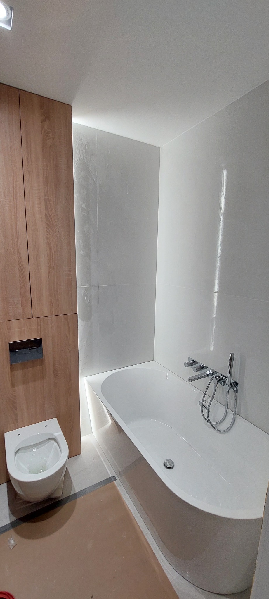 Nowoczesna łazienka z białą wanną, wiszącą toaletą i drewnianą zabudową, wykończona jasnymi płytkami z subtelną fakturą, oświetlona ledowym światłem.