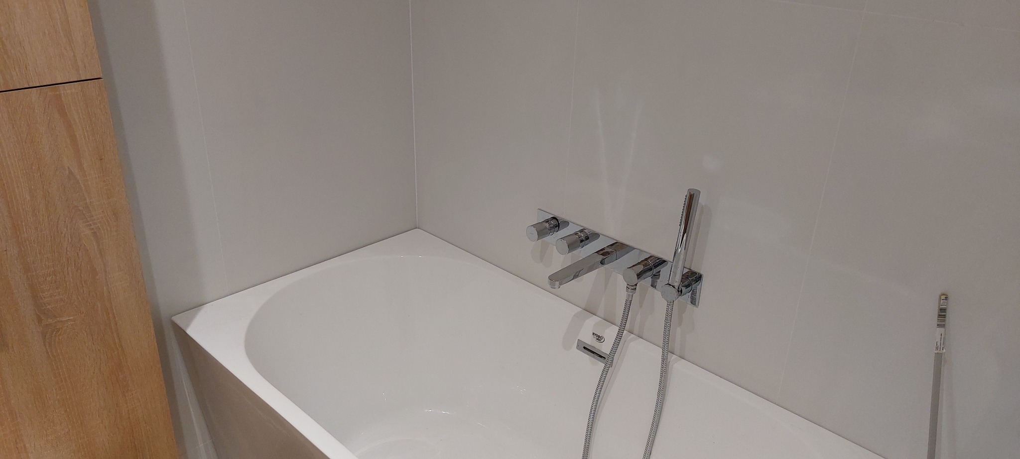 Biała wanna z chromowaną baterią ścienną, jasne płytki na ścianach, fragment drewnianej zabudowy z lewej strony.