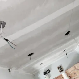 Sufit podwieszany w trakcie montażu z widocznymi otworami na oświetlenie i wystającymi przewodami elektrycznymi, szpachlowane łączenia płyt gipsowo-kartonowych.