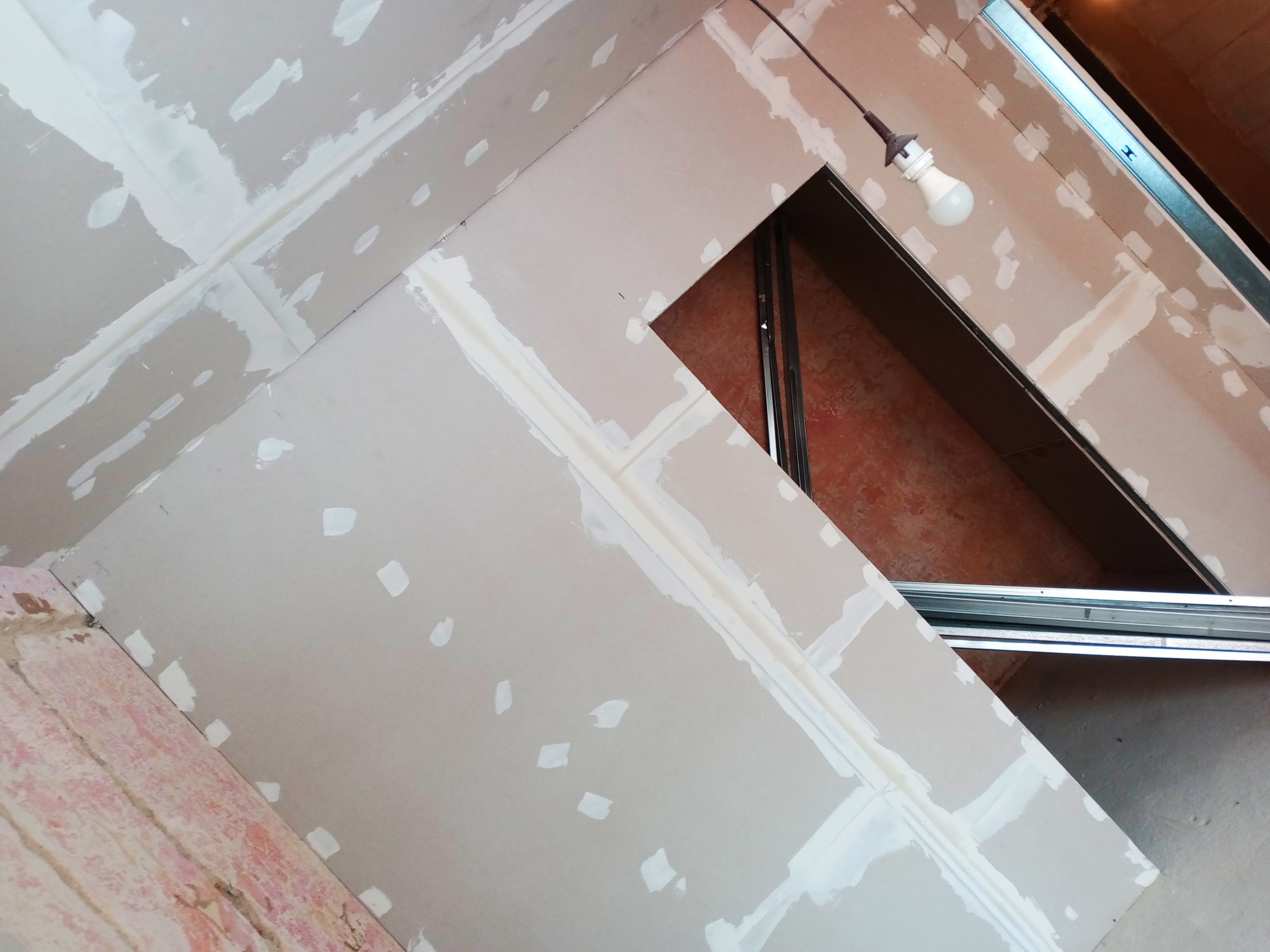 Skośny sufit poddasza w trakcie prac wykończeniowych, widoczne płyty gipsowo-kartonowe ze szpachlowanymi łączeniami i wkrętami, metalowy profil konstrukcyjny, żarówka na kablu oświetla wnętrze.