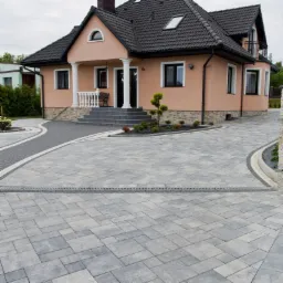 Szara kostka brukowa ułożona na podjeździe przed domem z kolumnami, widoczny system odprowadzania wody, ozdobne obrzeża i roślinność.