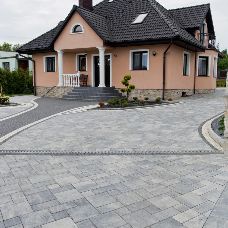 Szara kostka brukowa ułożona na podjeździe przed domem z kolumnami, widoczny system odprowadzania wody, ozdobne obrzeża i roślinność.