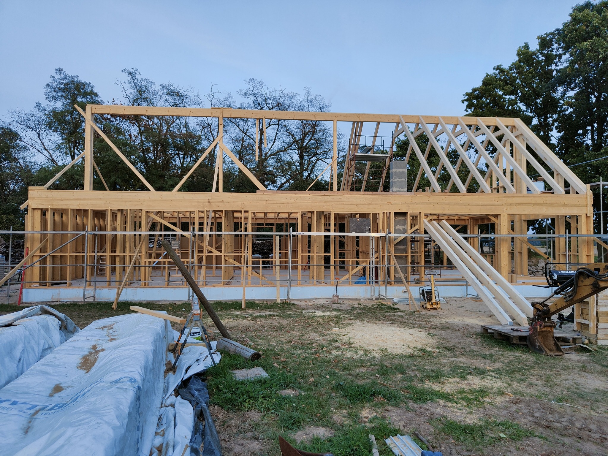 Konstrukcja szkieletowa domu jednorodzinnego w trakcie budowy, widoczne drewniane elementy nośne, rusztowanie, folia ochronna na materiałach budowlanych i sprzęt budowlany na działce.