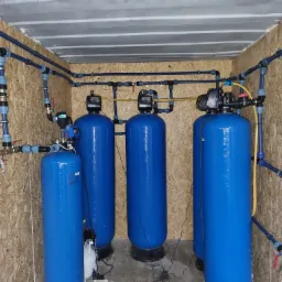 Kompletny system uzdatniania wody w pomieszczeniu wyłożonym płytami OSB, składający się z trzech niebieskich zbiorników ciśnieniowych z elektronicznymi sterownikami, połączonych niebieskimi rurami...