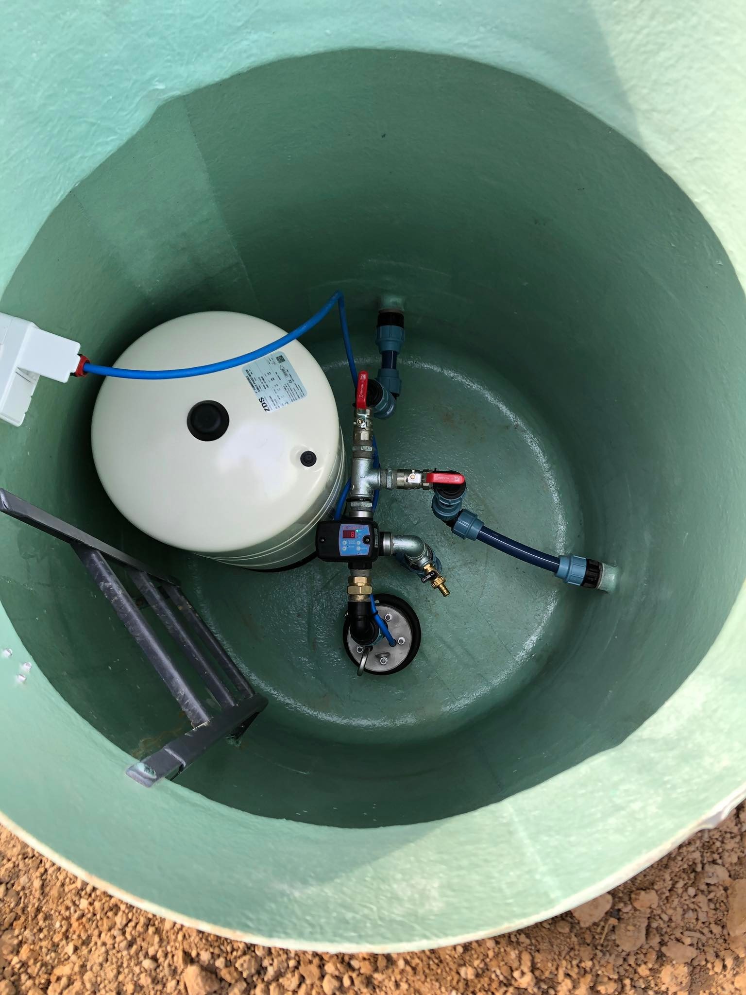 Wnętrze zielonej studni kręgowej z zainstalowanym zbiornikiem hydroforowym, rurami i armaturą, widok z góry.