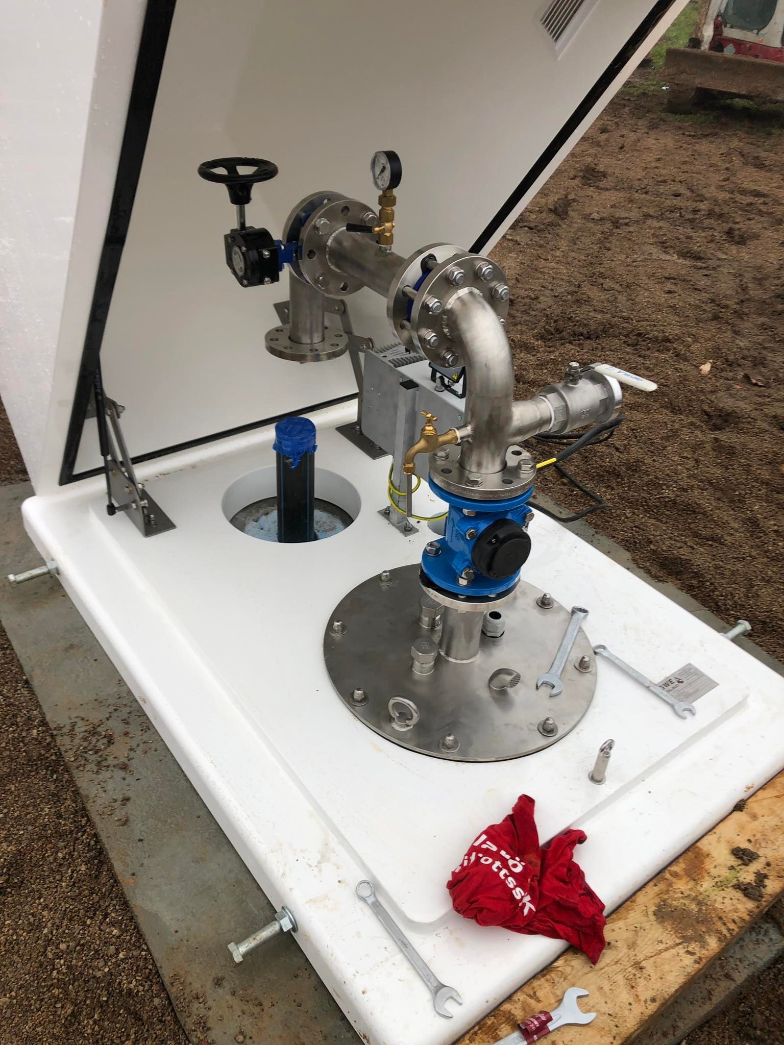 Gotowa instalacja studni w Kwidzynie, widoczne metalowe rury, zawory, manometr oraz czarna rura wystająca z otworu, całość zamknięta w białej skrzynce.