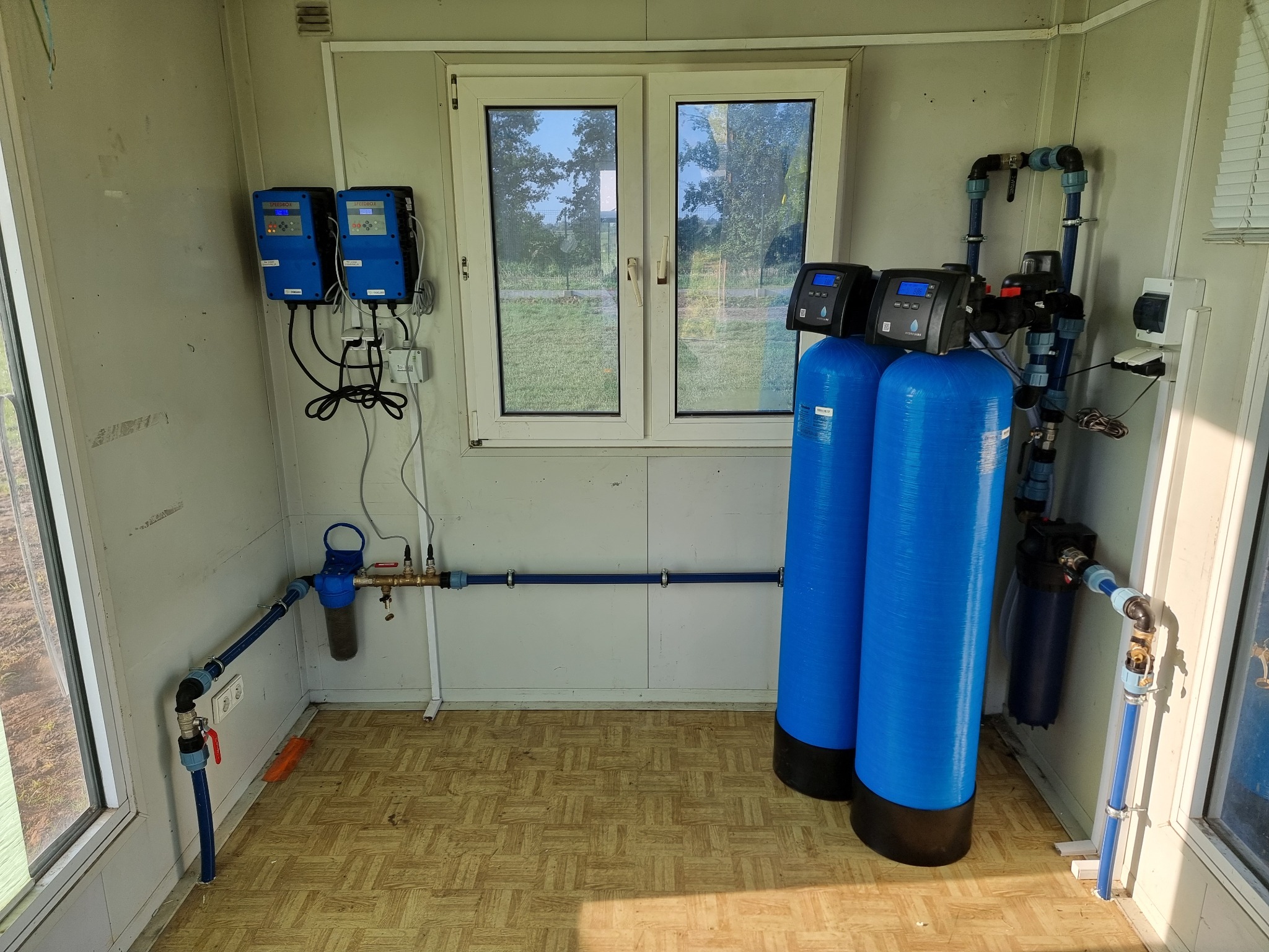 Wnętrze pomieszczenia z zainstalowanym systemem uzdatniania wody: dwa niebieskie zbiorniki ciśnieniowe z panelami sterowania, rury z zaworami i filtrami, kontrolery elektroniczne na ścianie, widok...
