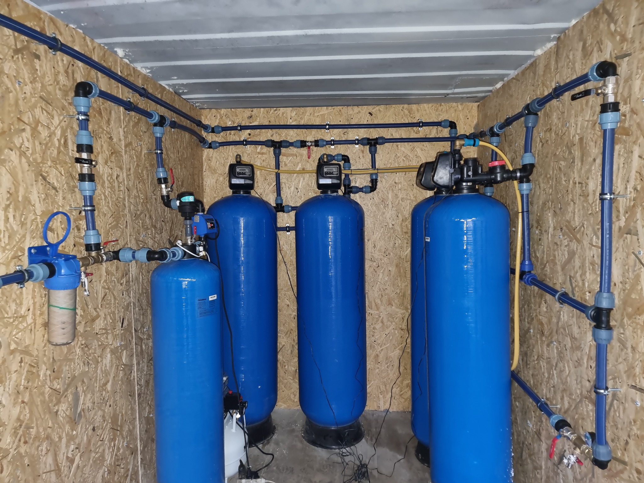 Kompletny system uzdatniania wody w pomieszczeniu wyłożonym płytami OSB, składający się z trzech niebieskich zbiorników ciśnieniowych z elektronicznymi sterownikami, połączonych niebieskimi rurami...