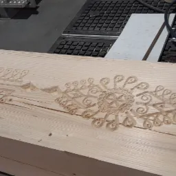 Drewniana belka z wyfrezowanym wzorem dekoracyjnym, widoczne ślady obróbki, w tle maszyna stolarska.