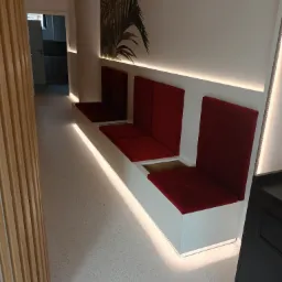 Długi, wbudowany siedzisko z czerwonymi poduszkami i oświetleniem LED, z otwartym schowkiem pod jednym z siedzeń, widok na kuchnię w tle.