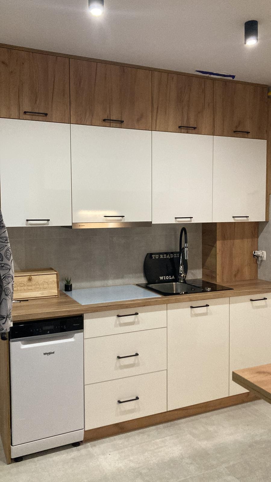 Nowoczesna kuchnia z białymi i drewnianymi szafkami, czarnymi uchwytami, szarą ścianą nad blatem i czarną armaturą. Na blacie drewniana skrzynka na chleb i mała roślina doniczkowa.