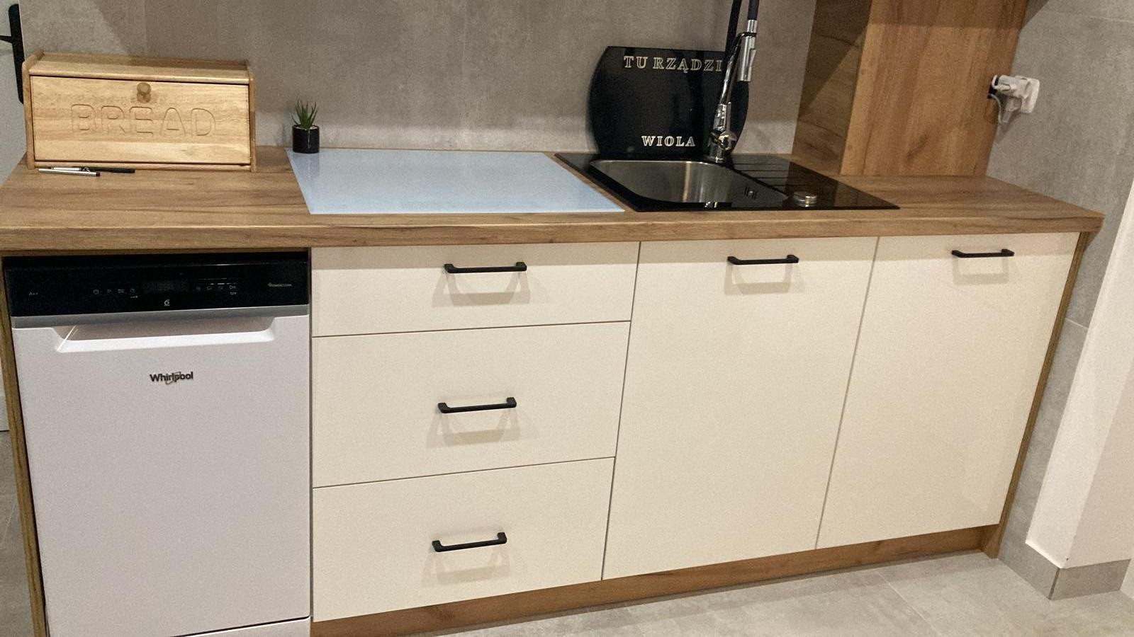 Biała zabudowa kuchenna z drewnianym blatem, czarnymi uchwytami i czarnym pojemnikiem na przybory kuchenne z napisem 'Tu rządzi Wiola'.