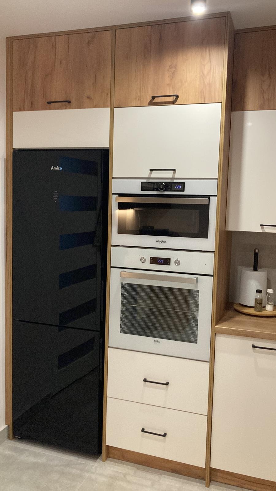 Nowoczesna zabudowa kuchenna z czarną lodówką Amica, białymi szafkami i drewnianymi akcentami, obejmująca mikrofalówkę Whirlpool i piekarnik Beko.