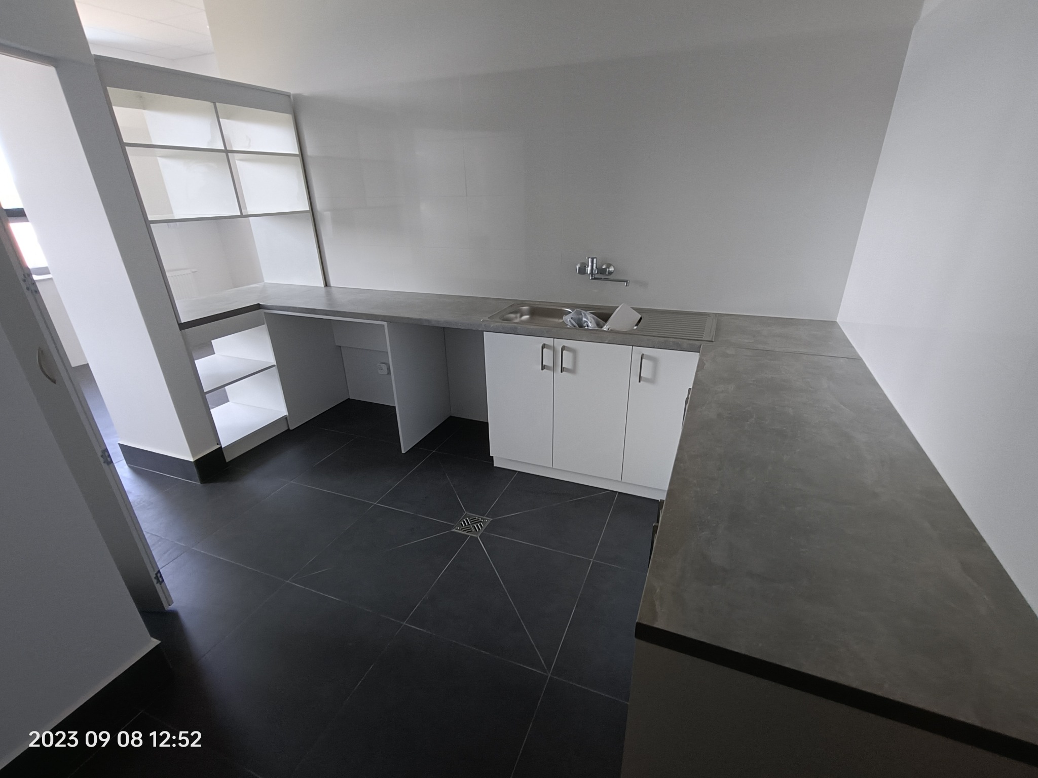 Wnętrze pomieszczenia z zabudową kuchenną z białymi szafkami i szarym blatem, widoczny zlew i bateria, czarna podłoga z płytek, w tle regał z półkami.