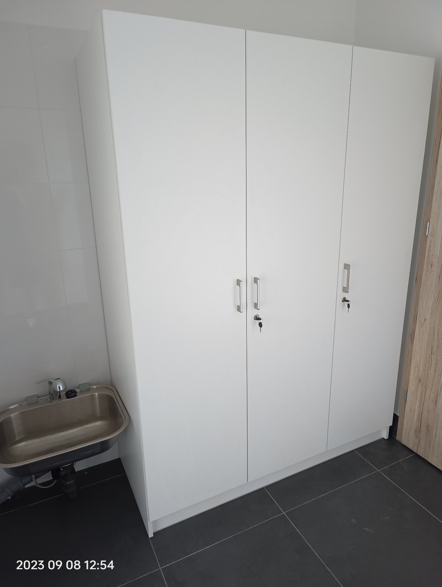 Biała szafa trzydrzwiowa z metalowymi uchwytami i kluczykami w zamkach, obok umywalki ze stali nierdzewnej w pomieszczeniu wyłożonym białymi płytkami i ciemnoszarą podłogą