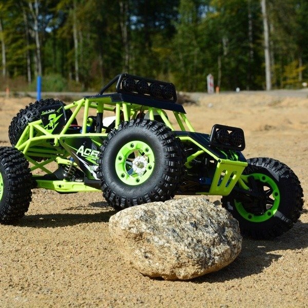 Zielono-czarny model samochodu terenowego RC z dużymi oponami, pokonujący przeszkodę w postaci kamienia na piaszczystym terenie, z lasem w tle.