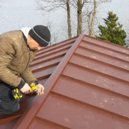 Monter w czapce, w trakcie montażu brązowej blachy trapezowej na dachu, w tle jezioro i drzewa.