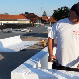 Pracownik w koszulce z logo 'MAZUR DACH' przycina nożycami blachę na dachu pokrytym papą, widoczne elementy obróbek blacharskich i narzędzia.