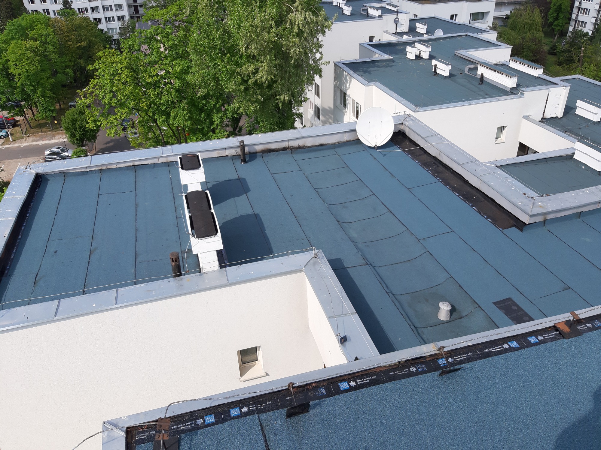 Widok z góry na płaski dach budynku wielorodzinnego pokryty papą termozgrzewalną, widoczne kominy wentylacyjne, antena satelitarna i detale obróbek blacharskich.
