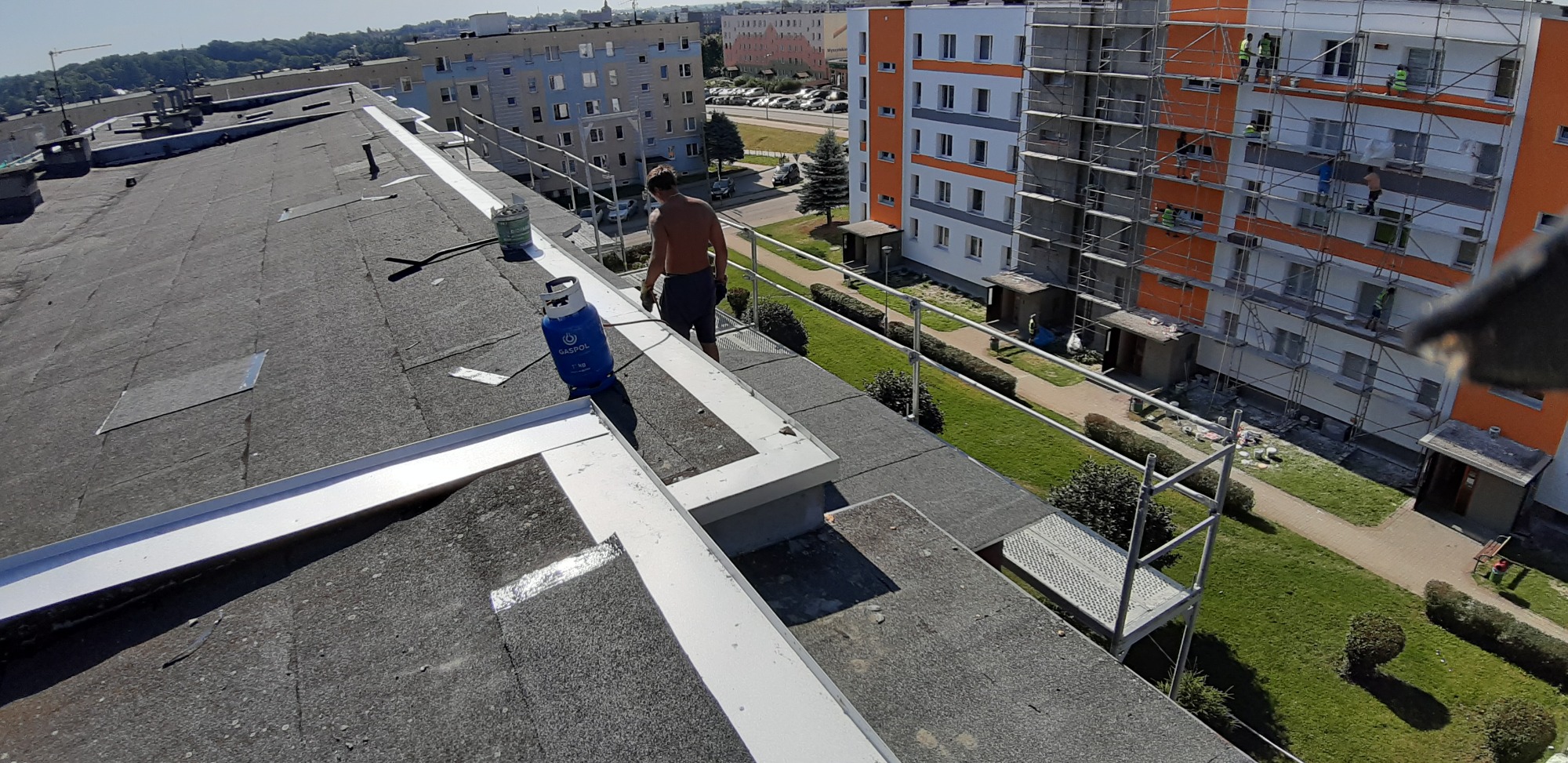 Prace dekarskie na dachu bloku w Boże, widoczny pracownik z butlą gazową i palnikiem, w tle budynek w rusztowaniach.