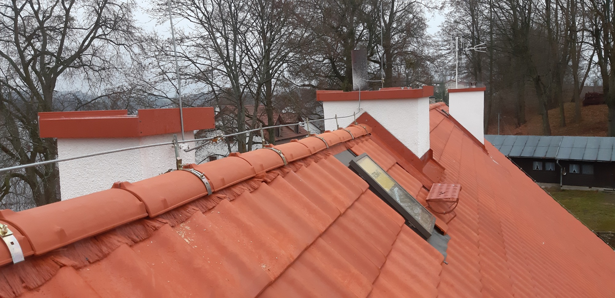 Czerwona dachówka ceramiczna na skośnym dachu z widocznymi elementami mocującymi, kominami w tle i fragmentem okna dachowego.