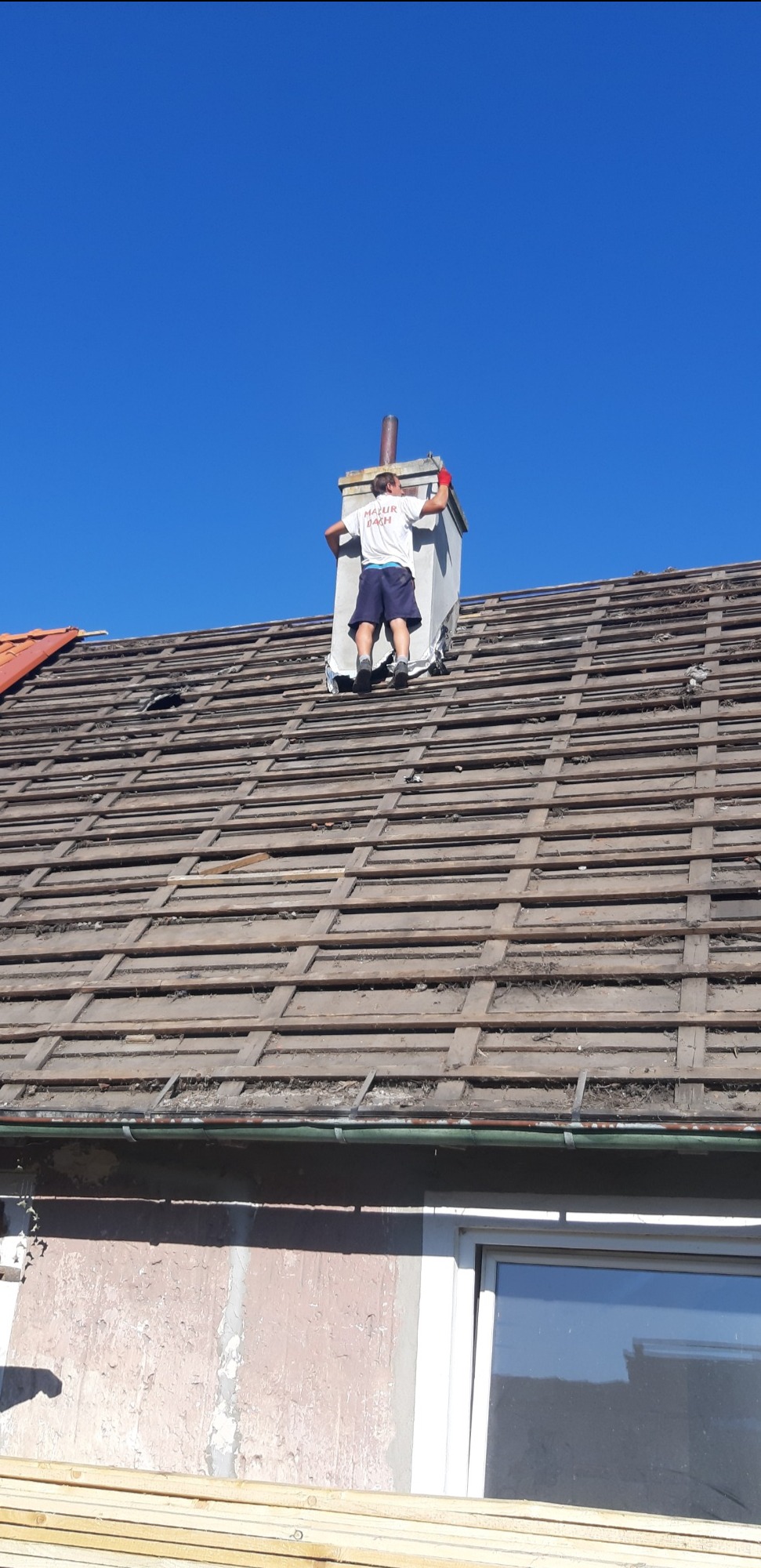 Pracownik w czerwonych rękawicach stoi na dachu z odsłoniętą konstrukcją więźby, przy kominie, podczas remontu dachu.