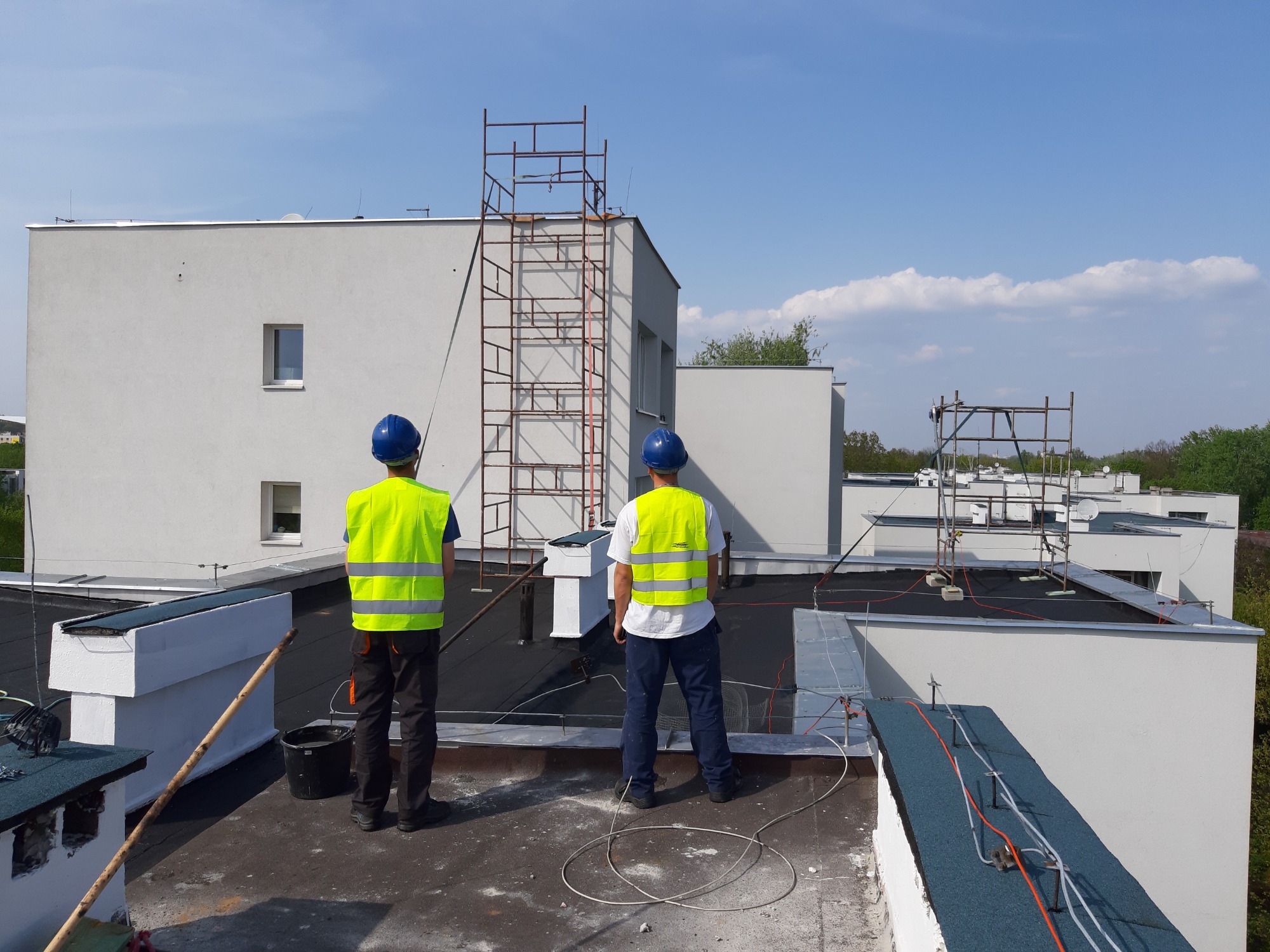Dwóch pracowników w kaskach i kamizelkach odblaskowych stoi na płaskim dachu budynku, widoczne elementy konstrukcji dachu, rusztowanie i sąsiednie budynki w tle.