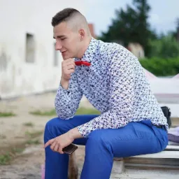 Młody mężczyzna w błękitnym garniturze i koszuli w kwiaty, z czerwoną muszką, siedzi zamyślony na ławce, trzymając telefon. Stylizacja na imprezę.