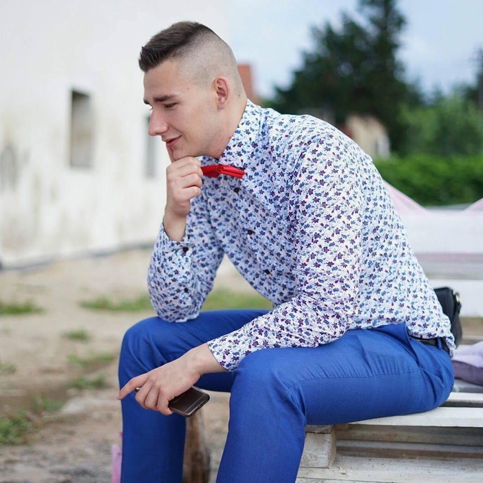 Młody mężczyzna w błękitnym garniturze i koszuli w kwiaty, z czerwoną muszką, siedzi zamyślony na ławce, trzymając telefon. Stylizacja na imprezę.