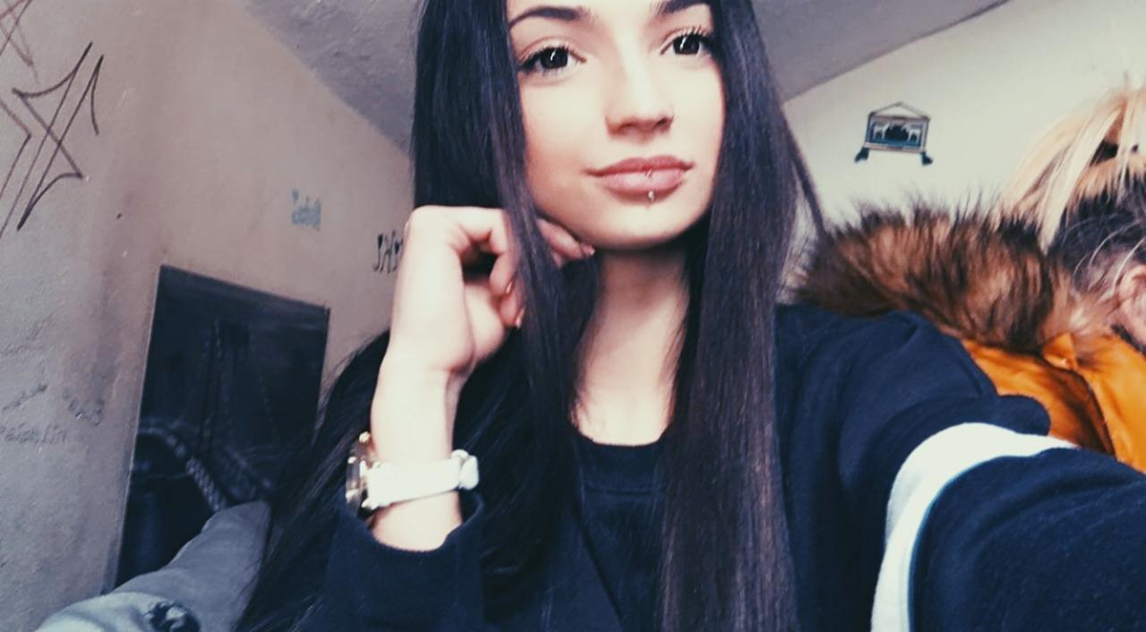 Portret młodej kobiety z piercingiem w wardze, opierającej brodę na dłoni. W tle ściana z graffiti i element kurtki z futrzanym kapturem.